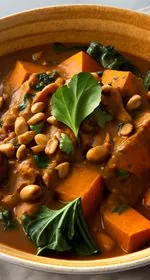 African peanut stew in an ornate white bowl sits in the center on a wooden cutting board. The stew is topped with strips of jasmine rice, chopped collard greens, and chopped peanuts. To the right of the bowl is a white and blue checkered tea towel. To the left of the bowl is half of a lime and sliced sweet potato. Scattered about are chopped collard greens and chopped peanuts. At the top of the image are the words 'The Panicked Foodie,' with the i's colored red and dotted with red hearts. At the end is a black mixing bowl with a red heart on it that contains three sugar cubes.
