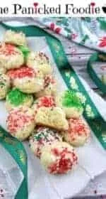Gluten-free ricotta cookies sit on a scalloped cream colored serving tray. On top of the pile is half of a cookie to show the fluffy interior texture. A green ribbon with gold leaves weaves throughout the photo. In the upper right is a Christmas themed tea towel and one more gluten-free ricotta cookie. Scattered throughout the photo are red and white sprinkles. Everything sits on a wood plank background. At the top of the image are the words 'The Panicked Foodie,' with the i's colored red and dotted with red hearts. At the end is a black mixing bowl with a red heart on it that contains three sugar cubes.